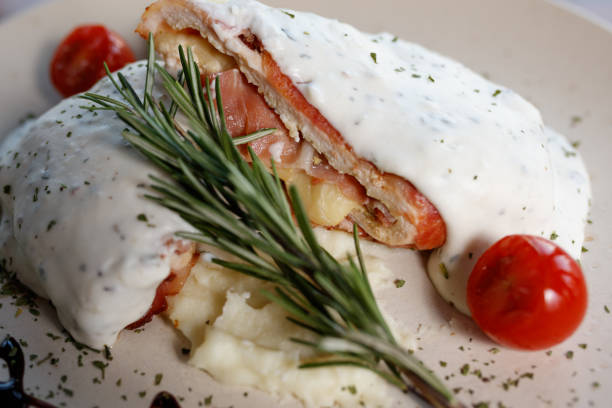 A close-up of a stuffed chicken breast topped with creamy sauce, garnished with rosemary and accompanied by cherry tomatoes and mashed potatoes, presented on a plate for fine dining enjoyment