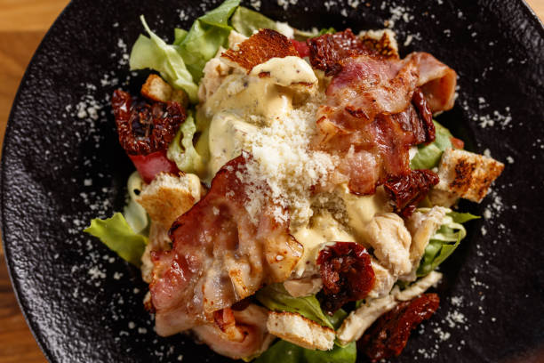 A close-up view of a Caesar Salad featuring crispy bacon, sun-dried tomatoes, and croutons. Served on a black plate, it highlights fresh ingredients for a savory and delicious dining experience