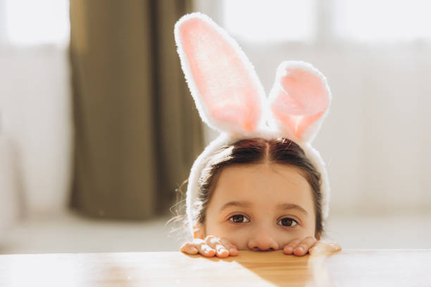 istock Happy Easter! Cute little girl wearing bunny ears while hiding behind table 2195823969