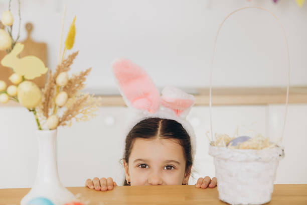 istock Happy Easter! Cute little girl wearing bunny ears while hiding behind table 2195823975