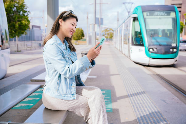 istock Smiling Young Woman Using Smartphone While Waiting for Tram at Station 2195844094
