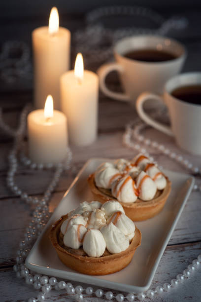 Two tarts topped with whipped cream and caramel drizzle on a white plate, surrounded by white candles, two white cups, and pearl necklaces on a wooden surface