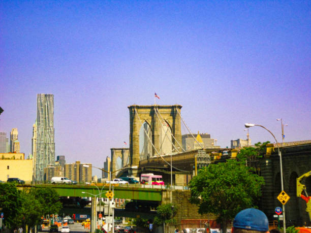 A colorful view of the iconic Brooklyn Bridge with the dynamic Downtown Manhattan skyline in the background. The bustling streets and elevated highways frame this quintessential New York City scene, blending historic architecture with modern skyscrapers. The vibrant blue sky adds a bright and energetic atmosphere to this urban landscape.