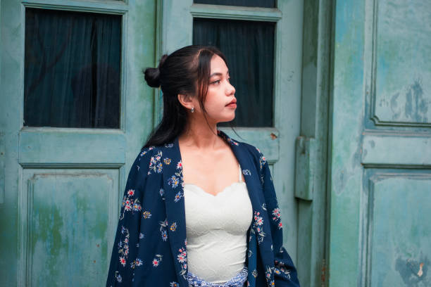 A young woman dressed in Javanese traditional attire poses gracefully at the doorway of an old Javanese house. The portrait beautifully showcases the harmony between her cultural clothing and the intricate design of the traditional architecture.