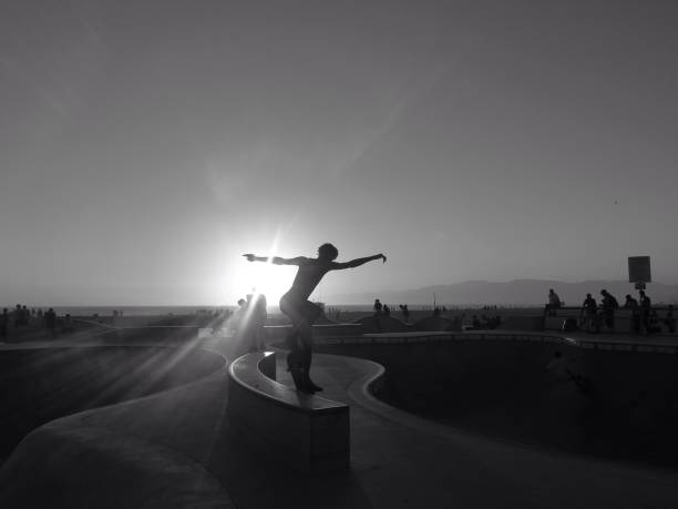 A low-angle view of a skateboarder poised on their board against the setting sun. Urban surroundings and distant figures accentuate the energetic and recreational vibe of skateboarding in public areas.