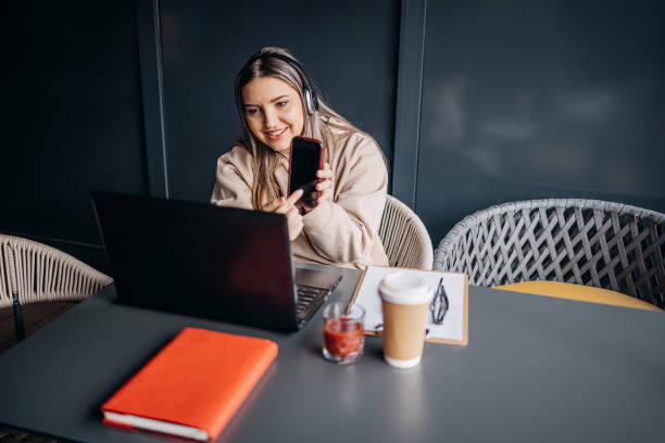 istock Young woman having drink alone at cafe and enjoying it while on a video call using laptop and showing something on mobile phone 2195914701