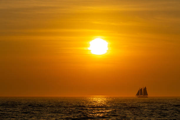The sun is setting on the horizon while a small sailboat is crossing the frame.