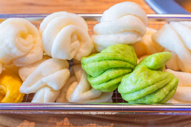 This image displays a delightful assortment of Chinese steamed buns, known as baozi, presented in a clear glass dish. The collection includes classic white baozi, which are plump and feature a delicate, pleated design, showcasing the skill involved in their creation. Alongside the white baozi are vibrant green leaf-shaped buns, likely flavored with pandan or spinach, adding a touch of color and variety to the assortment. The buns are arranged neatly, allowing each one to be visible, and their glossy surface indicates they are freshly steamed and ready to be enjoyed. This image captures the essence of Chinese dim sum culture, where such buns are a popular choice for their delicious taste and the artistry of their presentation.
