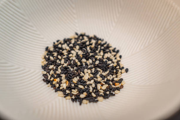 The image shows a close-up of a pile of black and white sesame seeds in a light-colored bowl. The sesame seeds are scattered in the center of the bowl, forming a small mound. The contrast between the dark black seeds and the light white seeds creates a visually appealing pattern. The bowl appears to be made of ceramic or porcelain, with a smooth, slightly textured surface. The background is blurred, focusing attention on the sesame seeds. The image is simple yet elegant, highlighting the natural beauty and texture of the sesame seeds.