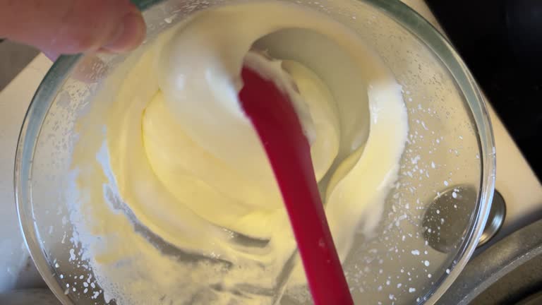 A stainless steel bowl sits on a kitchen bench, holding a swirl of cake mix as a light fluffy merangue is, stirred gently with a spatula