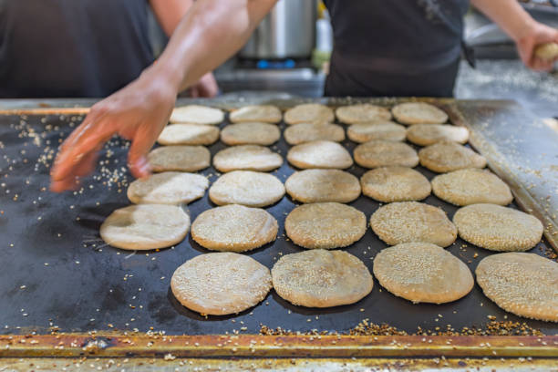 This image captures the skilled hands of a baker at work, preparing sesame huoshao, a beloved Northern Chinese pastry. The huoshao, which are round and generously sprinkled with sesame seeds, are neatly arranged on a large, dark baking tray. The baker's hand is seen sprinkling seeds onto the dough, a crucial step that not only adds flavor but also gives the huoshao their distinctive texture. The tray is already populated with numerous huoshao, some of which are golden-brown, indicating they are nearly ready for the oven. The background, though blurred, suggests a bustling kitchen environment where these pastries are being made in large quantities, likely for a market or a bakery. The image is a testament to the art of traditional baking, highlighting the meticulous process and the hands-on craft that goes into making each sesame huoshao.