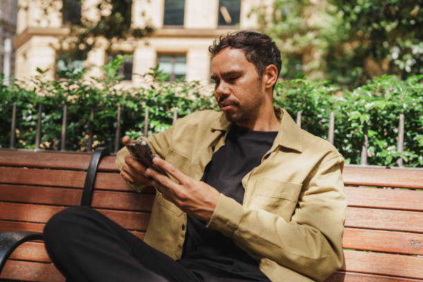 istock Man using his smartphone while seated on a wooden bench in a city park 2195989028
