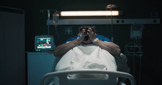 istock Exhausted tired man with headache suffering from migraine holding head with hands sitting on hospital bed 2195993059