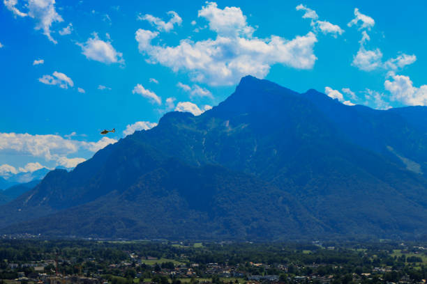 A panoramic view of Salzburg with lush greenery, scattered houses, and towering mountains in the distance under a bright blue sky with scattered clouds.