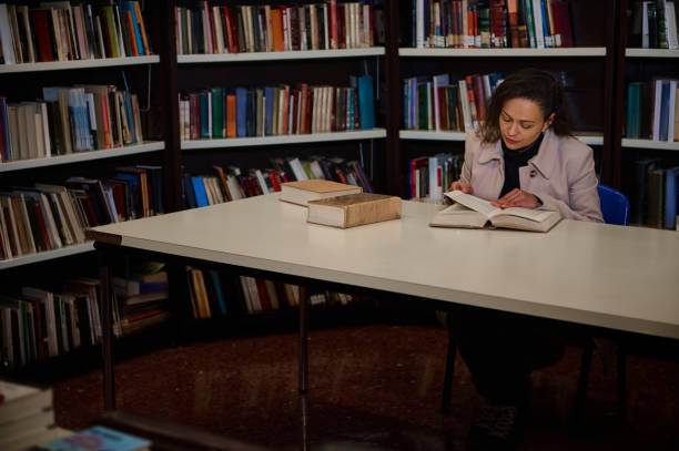 Woman reading a book alone at a library table among bookshelves