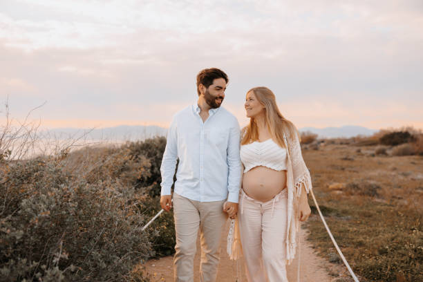 Joyful young pregnant couple walking hand in hand along a path between the dunes sharing loving smiles as the pastel hues of a sunset glow behind them