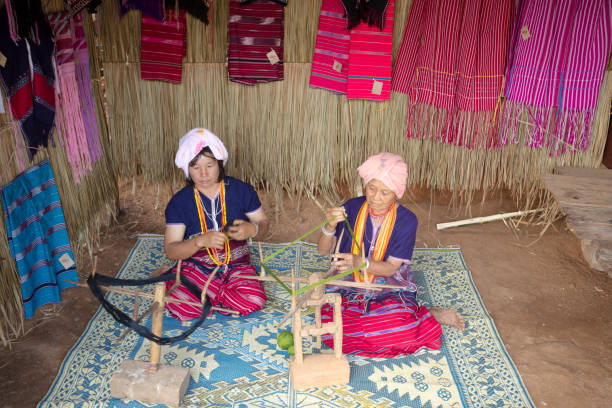 Two thai hill tribe women are rolling up wool for looming.  Women are wearing headscarf and traditional clothes.  They are sitting below stilt house in rural mountain town Pangkhum in Amphoe Samoeng in eastern Chiang Mai province. Women are sitting on ground and plaids. In front of them are spinning tools for wool. Women are wearing local traditional clothes. \n In background some clothes and textiles examples are hanging at wooden wall. View from position in front of the house.