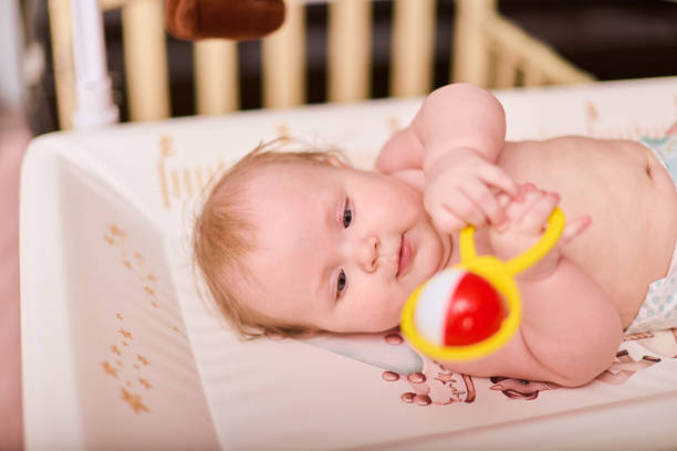 Baby girl with blonde hair holding colorful toy on changing table. Soft lighting enhances gentle expression. Wooden crib in softly blurred background