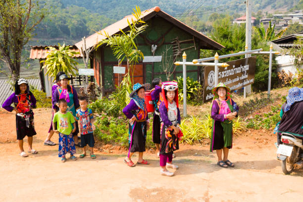 Traditionally dressed thai women in mountains are standing near and in front of former old school building of town. Scene is in mountain town Pangkhum in Amphoe Samoeng in eastern Chiang Mai province. There are adult and mature women. One woman is holding a bottle of water and is smiling into camera. Among women are  a few children in left area. A person is passing by on  a motorcycle at right side. In background are mountains and in top right area are houses of town. In front of small building is a sign with official thai authority logo and some  text in thai language.