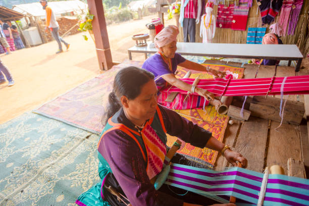 Two mature thai are looming in mountain town Pangkhum in Amphoe Samoeng in eastern Chiang Mai province. Women are sitting below stilt house. They are sitting on ground and plaids. Women are wearing local traditional clothes. They are looming different colored textiles. In background some clothes and textiles examples are hanging at wooden wall. In left area is open area in front and between houses . Some people are walking or standing there.
