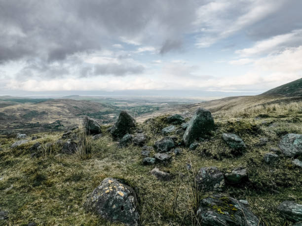 Hilltop scene. Several large rocks sit atop grassy knoll overlooking a wide valley. Cloudy sky.