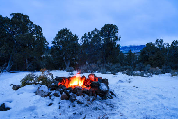 Campfire Burning in Fire Ring in Winter with Snow - Dusk blue hour with flames and heat in cold mountain environment.  Blue hour with glowing fire at campsite. Rock fire ring and orange flames.