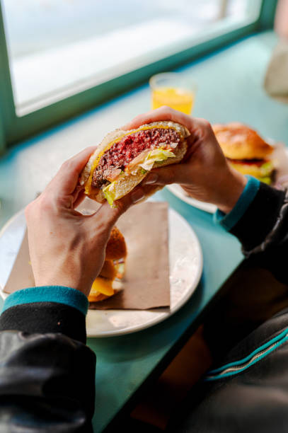 Hands holding a juicy burger with cheese, meat, and salad, next to a glass of orange juice in a restaurant