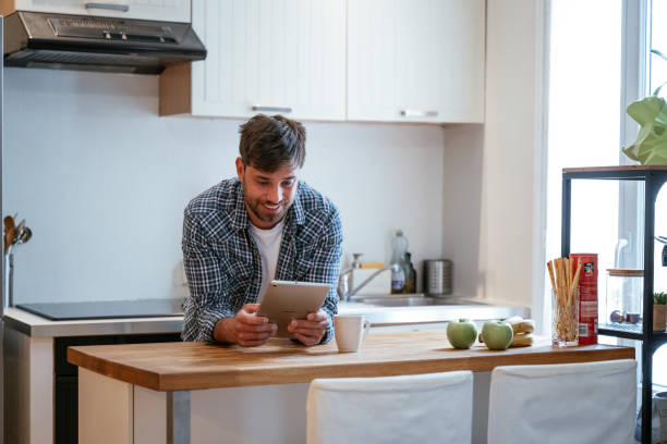 istock Young businessman in casual wear leaning on kitchen counter while looking at digital tablet 2196080022