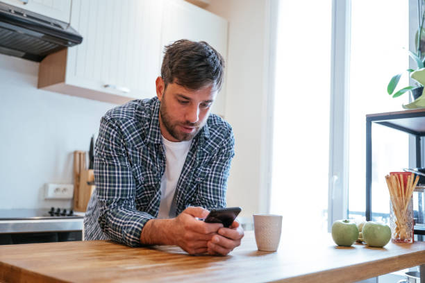 istock Serious young man in casual wear leaning on kitchen counter while looking at smartphone 2196080067