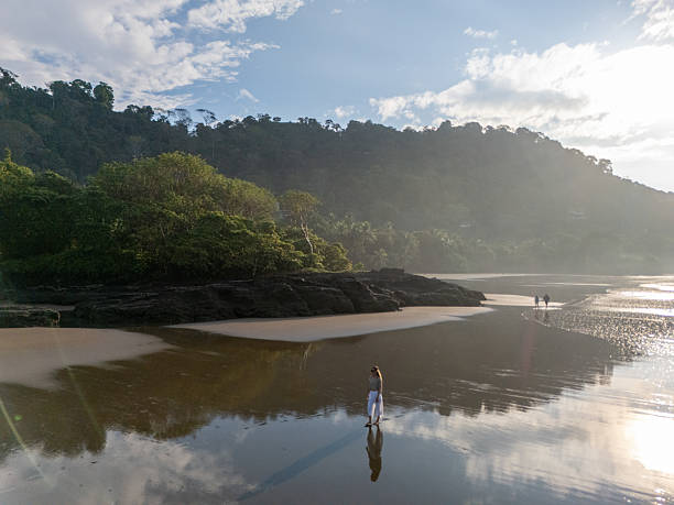 Woman walking along reflective beach, enjoying early morning solitude. Calm ocean landscape with moving figures and distant hills.