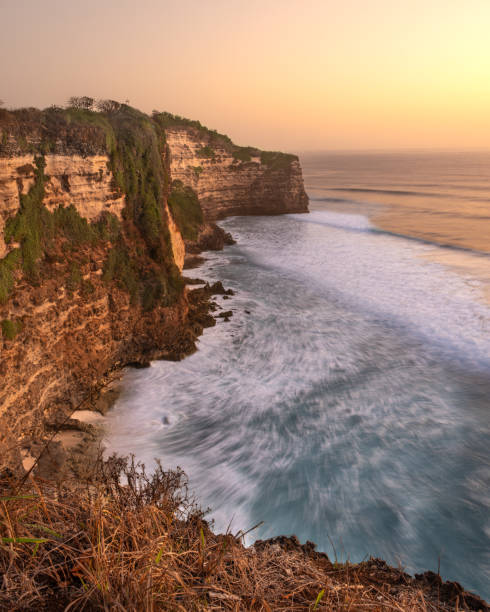 Golden light bathes the towering cliffs overlooking a restless ocean, where waves crash rhythmically against the rugged shoreline. Verdant greenery clings to the rock face, blending with the soft hues of a peaceful sunset. This stunning coastal landscape captures the harmony of nature’s raw power and serene beauty