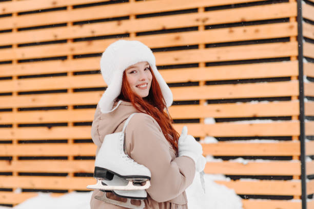young pretty caucasian redhead smiling girl in white and beige winter clothes skating in ice rink in winter cloudy snowy day, with skates on shoulder, looking straight to camera