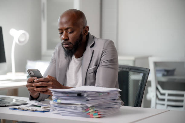 istock Businessman sitting at his desk using a smartphone with a large stack of paperwork in front of him 2196120920
