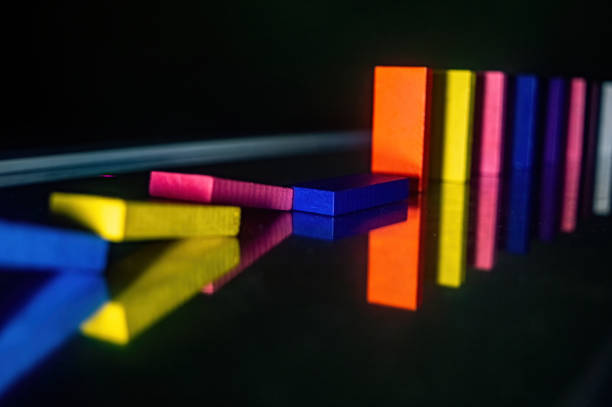 Colorful dominoes in a line, with some fallen over, reflecting on a shiny surface. The vibrant colors and reflections create a visually striking and dynamic composition