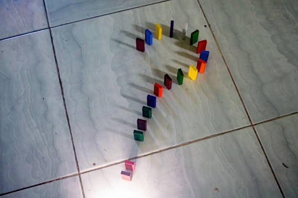 Colorful dominoes arranged in a question mark shape on a tiled floor, casting shadows. The arrangement and lighting create an intriguing visual effect