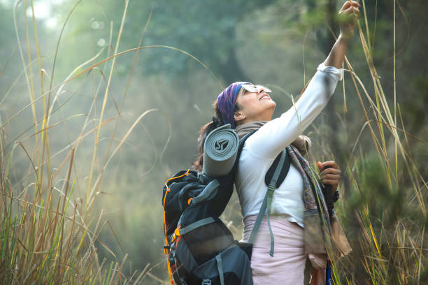 Side view of smiling young woman with backpack and camping mat touching plant while hiking in forest during weekend