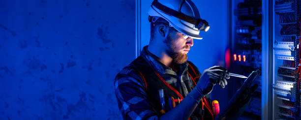 Electrician focused on work in switchboard with fuses, using tablet. Electrical engineer in special clothes with flashlight on helmet in dark room with emergency lights in background. Space for text.