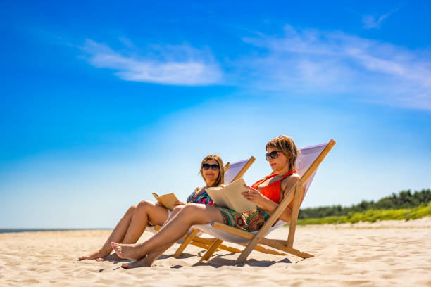 istock Two mature women reading books sitting on deckchairs on beach 2196133334