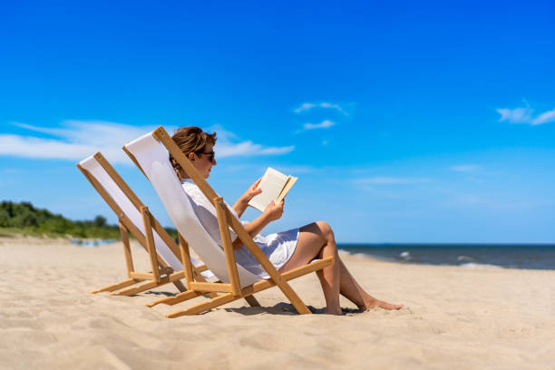 istock Mature women reading books sitting on deckchairs on beach 2196134448