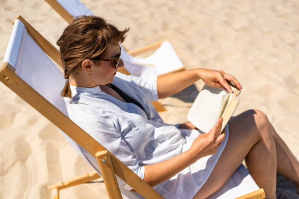 istock Mature women reading books sitting on deckchairs on beach 2196134464