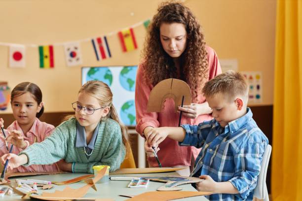 istock Kind Teacher Assisting Kids during Art Craft Class Painting Cardboard Figures 2196136677