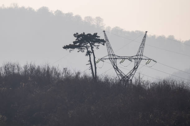 A high-voltage power line tower standing amidst a misty suburban landscape. The hazy background includes houses and hills, blending nature and infrastructure in a serene setting.