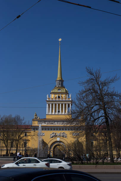 A wide-angle view of the iconic Admiralty Building in St. Petersburg, Russia. The building's golden spire and clock tower dominate the skyline against a clear blue sky. Cars and pedestrians add a sense of life and scale to the scene.
