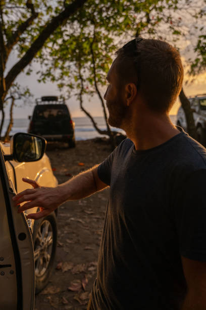 istock Man Standing Beside Open Car Door at Scenic Beachside Sunset 2196165179
