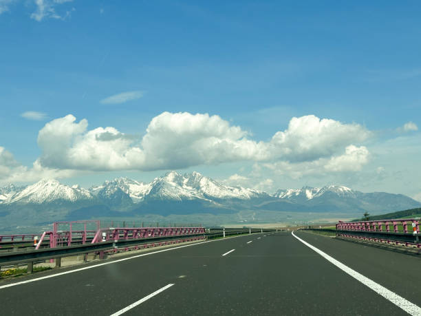 Wide highway leading through green fields towards majestic snow-covered mountain peaks Tatry under a bright blue sky with fluffy white clouds.  Tatry, Slovakia.