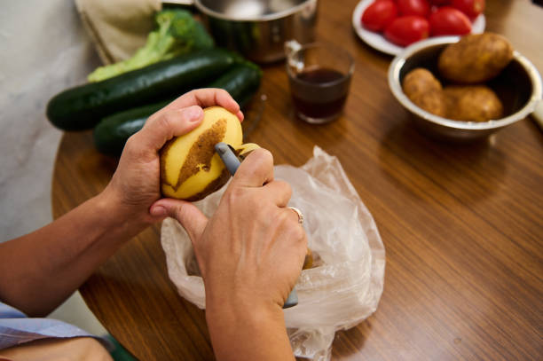 A close-up view of someone peeling a potato with zucchini, tomatoes, and utensils nearby.