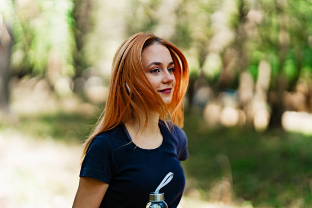 Beautiful young redhead woman walking and jogging in the woods with bottle of drinking water. Healthy lifestyle and sport in nature concept.