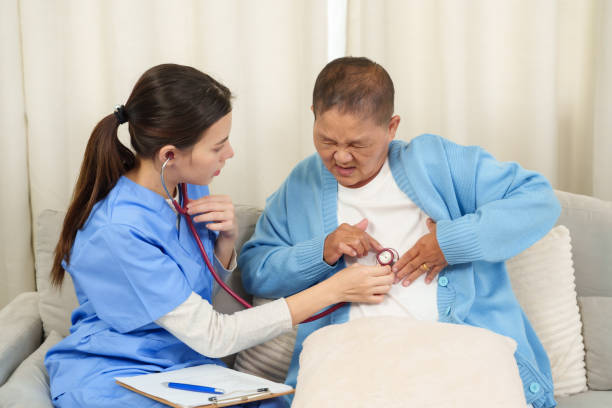 Nurse performing chest examination with stethoscope on elderly Asian woman experiencing discomfort during recovery. Home healthcare session providing focused and compassionate care.