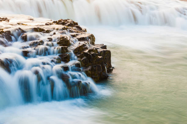 Water cascades gracefully over dark, rugged rocks, creating a serene atmosphere. The soft light of sunset enhances the natural beauty of this Icelandic waterfall, inviting tranquility.