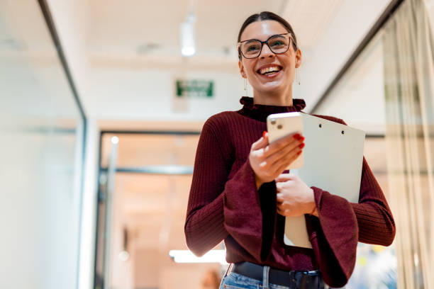 istock Businesswoman walking in office corridor using phone and holding clipboard 2196235501